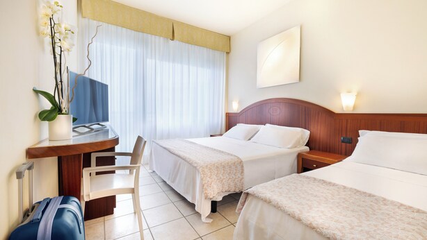 A hotel room with two beds, a desk with a chair, a blue suitcase, and a potted orchid. - Hotel Bellevue Beach (Milano Marittima)