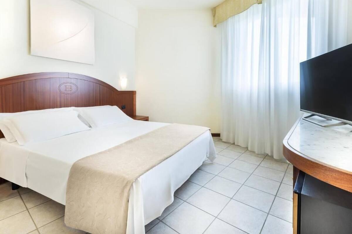 A hotel room with a bed, a TV, a nightstand, and a large window with curtains.