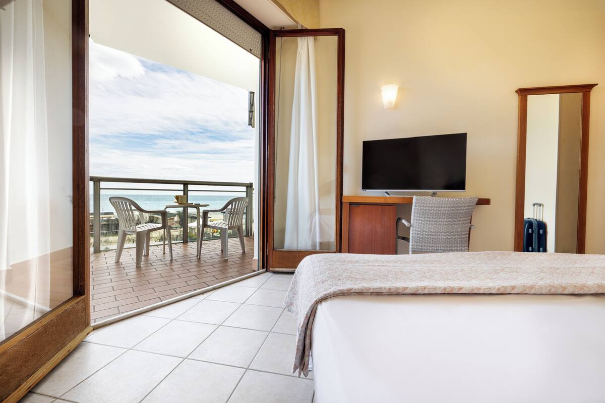 A hotel room with a bed, a desk with a TV, a chair, a suitcase, and a balcony with a view of the sea.