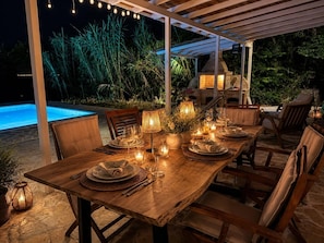 An outdoor dining area with a wooden table set for a meal, surrounded by chairs, and a pool in the background.