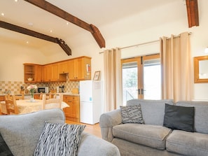 A living room with a grey sofa, a dining table with chairs, wooden cabinets, a kitchen area, and a large window with curtains.
