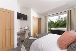 Ein Schlafzimmer mit Bett, Schreibtisch, Fernseher und einer Glastür zu einem Balkon mit Whirlpool.