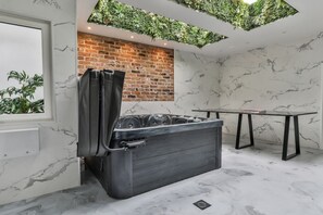 Un baño moderno con piso de mármol, una bañera negra, pared de ladrillo y un jardín vertical verde en el techo.