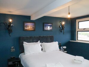 Un dormitorio con cama, dos luces empotradas en la pared, dos cuadros enmarcados, una ventana y una mesita de noche con lámpara.