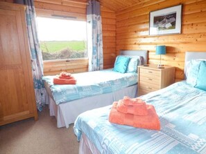 A room with two beds, a wooden ceiling, a window with curtains, a bedside table with a lamp, and a framed picture on the wall. - Charming Ty Pren Retreat in St Davids (Haverfordwest)