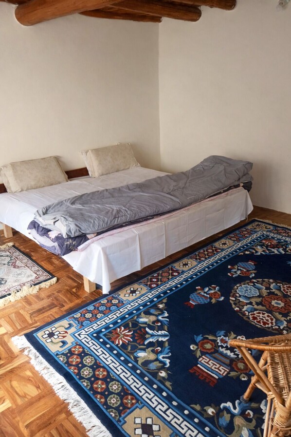 Una cama individual con colchón blanco y manta gris, un tapete estampado y una silla de mimbre.