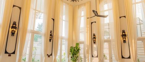 A spacious living room with a ceiling fan, chandelier, and large windows with curtains.