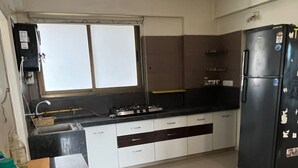 Cozinha com geladeira preta, armários brancos, janela com persianas e piso ladrilhado.