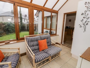 Une pièce comprenant un canapé en osier, un fauteuil, une table à manger et une grande fenêtre donnant sur un jardin et des maisons.