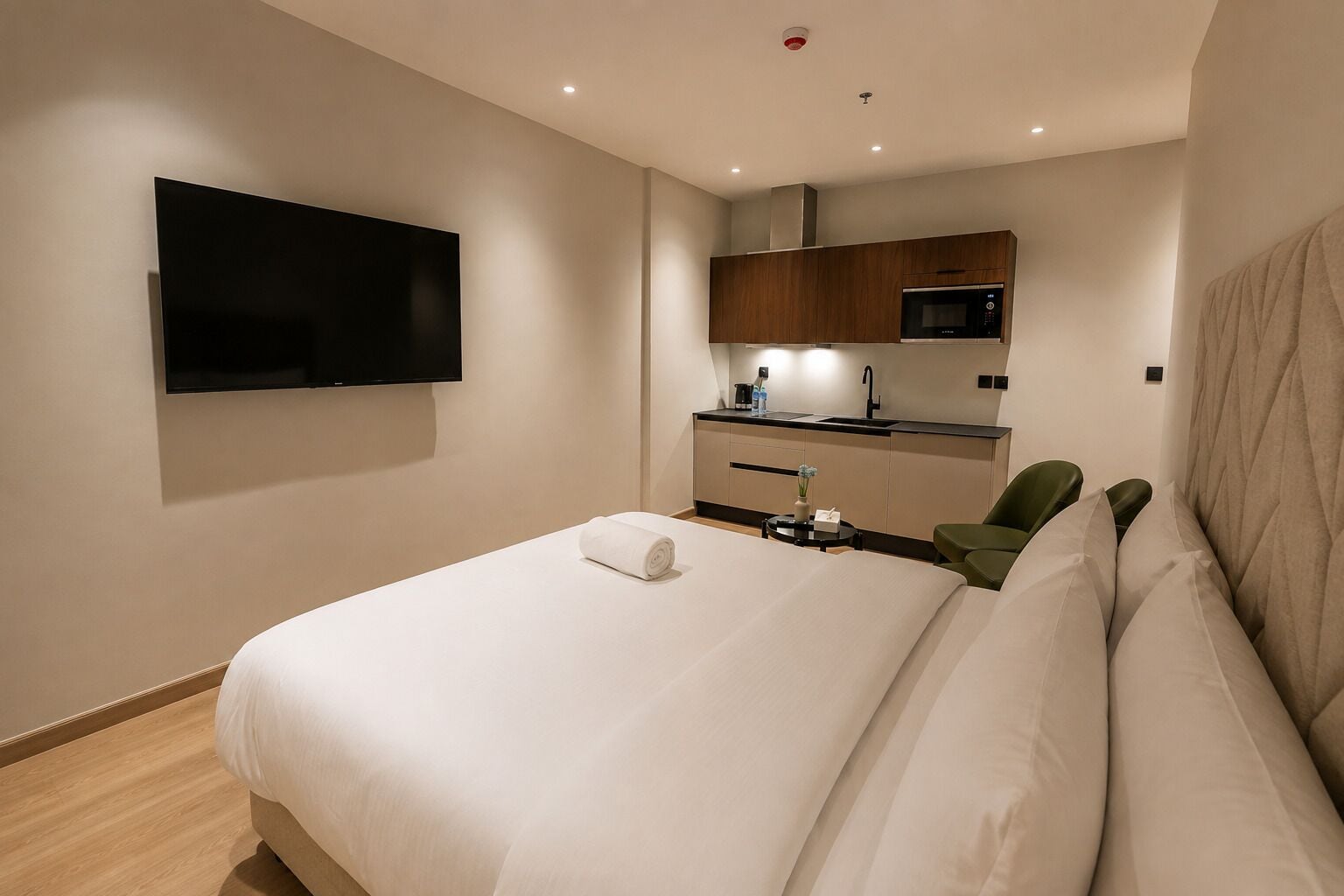 A hotel room with a bed, a flat-screen TV mounted on the wall, a kitchenette with a microwave, and a small table with a vase.