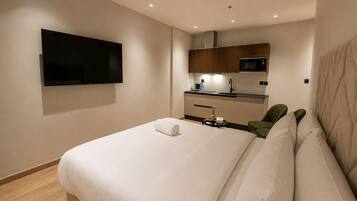 A hotel room with a bed, a flat-screen TV mounted on the wall, a kitchenette with a microwave, and a small table with a vase.