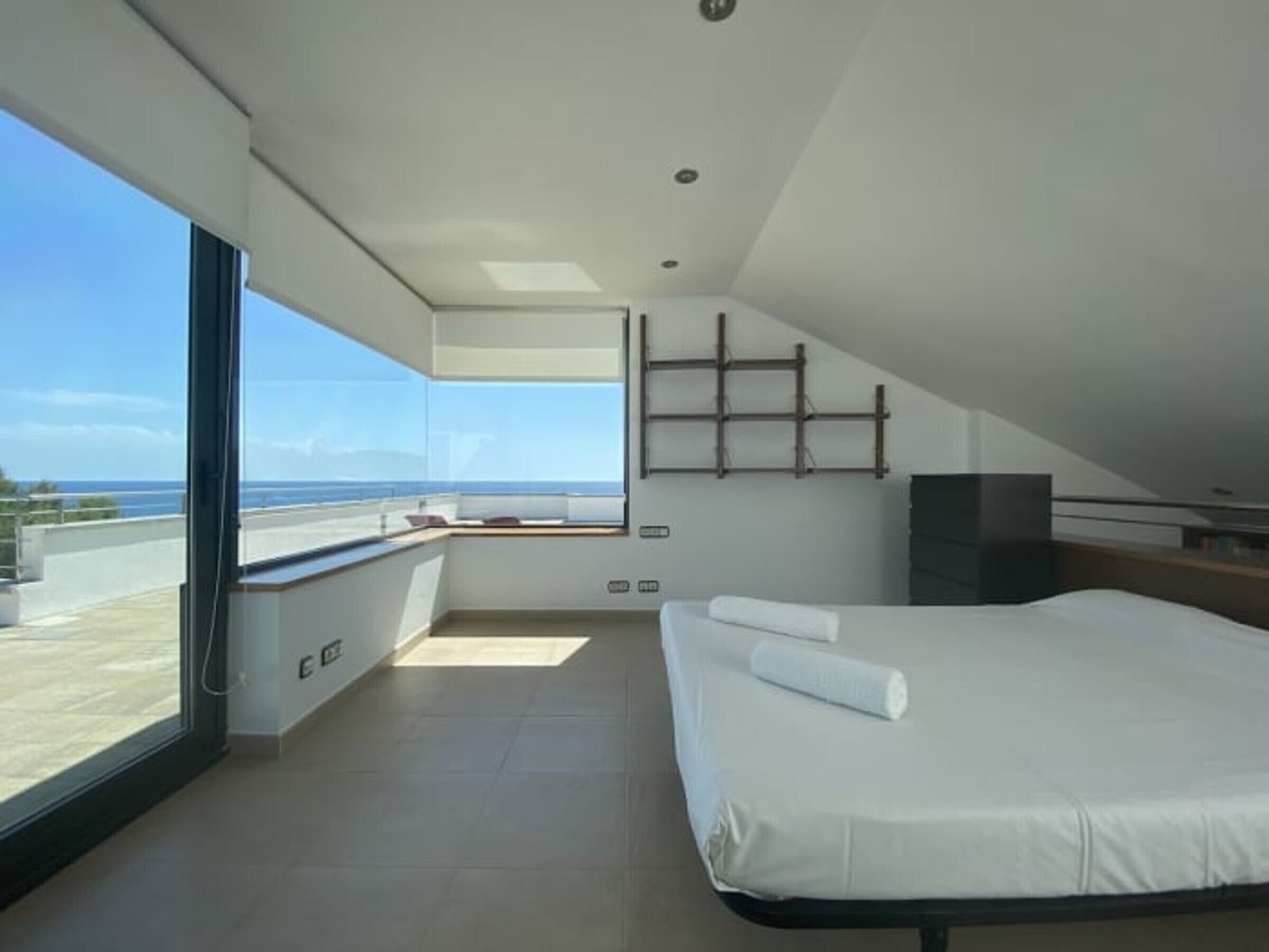 A modern bedroom with a large bed, a TV, and a balcony with a view of the sea.