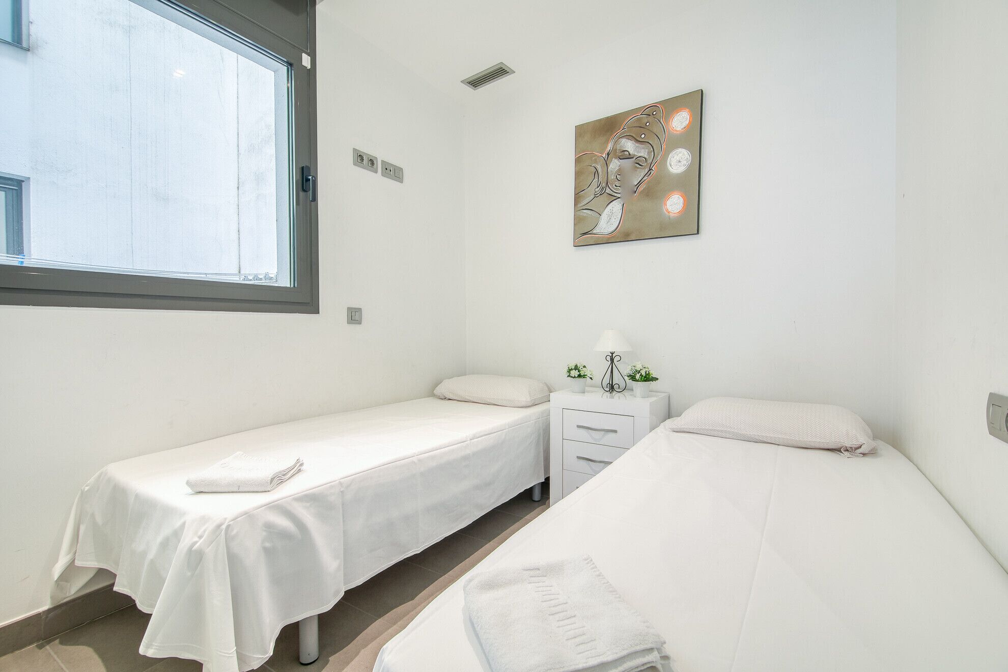 Una habitación con dos camas individuales, una ventana, una obra de arte en la pared y una mesita de noche con una lámpara y flores.