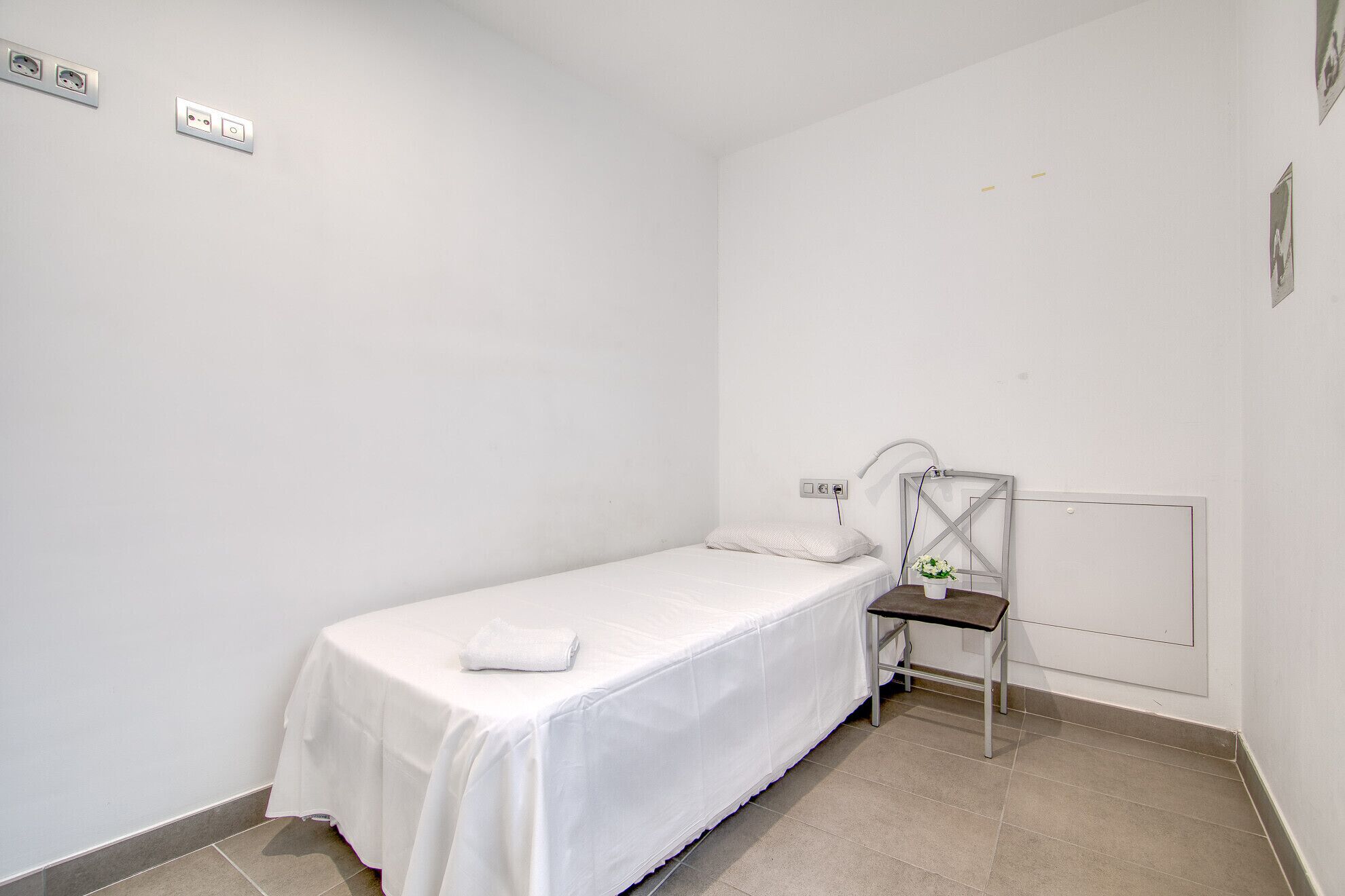 Une petite chambre avec un lit simple, une couette blanche, une table de chevet avec une lampe et une chaise.