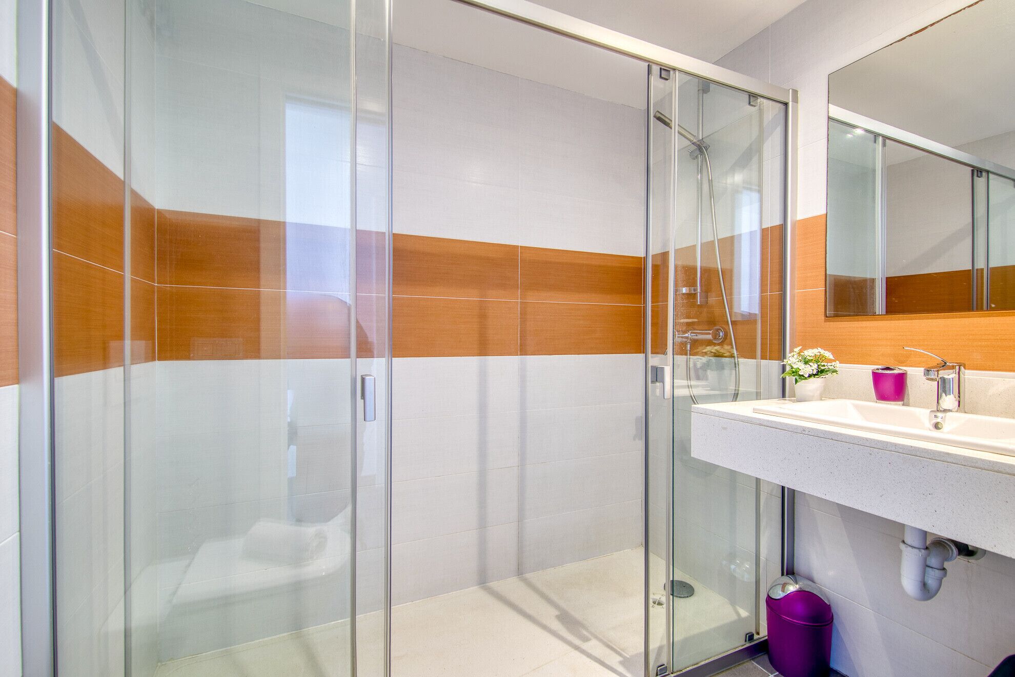 Une salle de bain moderne avec une douche à parois de verre, un lavabo blanc avec un miroir et une poubelle violette.