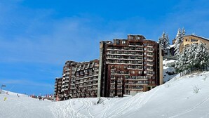 Ein Skigebiet mit einem großen Gebäude mit mehreren Balkonen, einer verschneiten Piste und einem klaren, blauen Himmel.