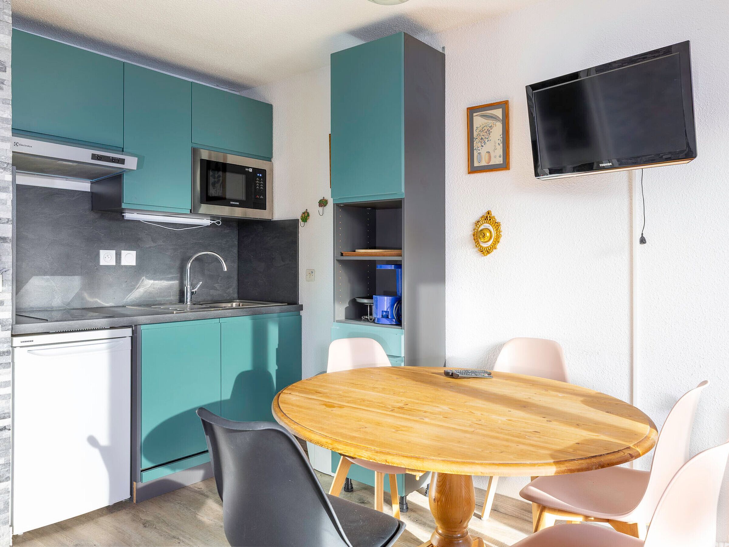 A compact kitchen with teal cabinets, a microwave, and a built-in oven. There is a round wooden dining table with chairs and a flat-screen TV mounted on the wall.
