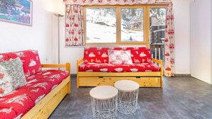 Een gezellige kamer met een houten bank, een rode gestreepte bank, twee ronde tafels en een groot raam met uitzicht op een besneeerd landschap.