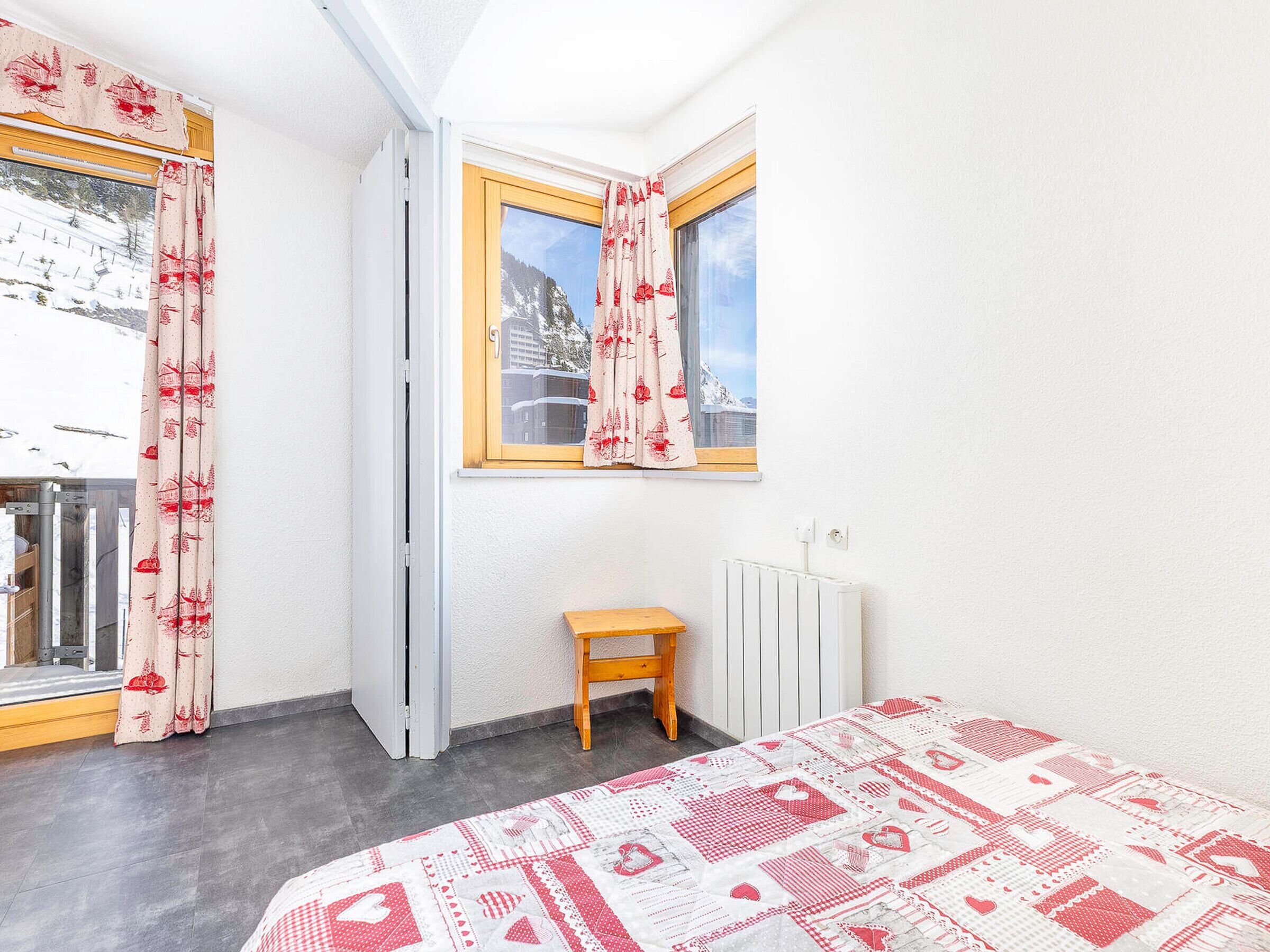 Une pièce comprenant un lit, un petit tabouret en bois, un radiateur et une fenêtre avec des rideaux.