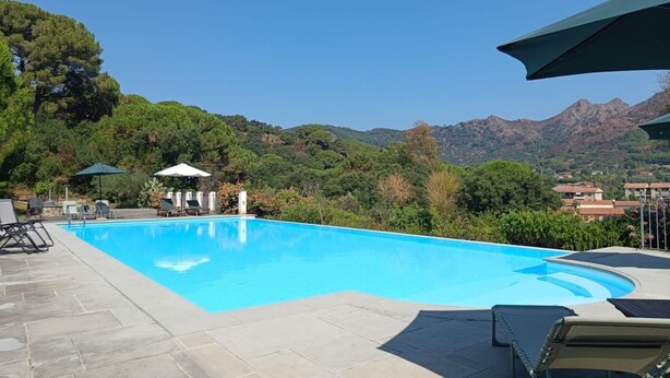 Une piscine d’un bleu éclatant, bordée de chaises longues et d’ombrelles, et entourée d’une végétation luxuriante et de montagnes.