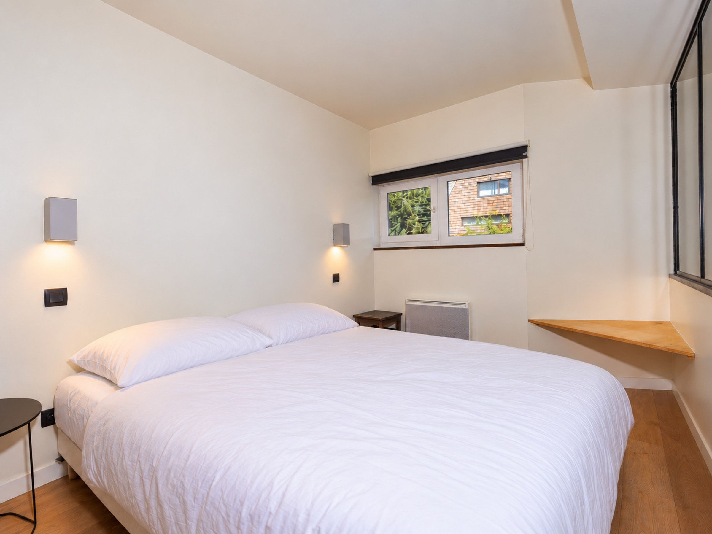 A bedroom with a large bed, two wall-mounted light fixtures, a radiator, and a window with a view of greenery.