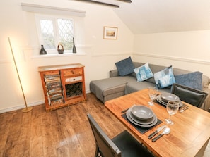 A dining area with a wooden table set for two, a sofa with cushions, a small cabinet with drawers, and a window with decorative vases. - The Hayloft, St Florence (Tenby)