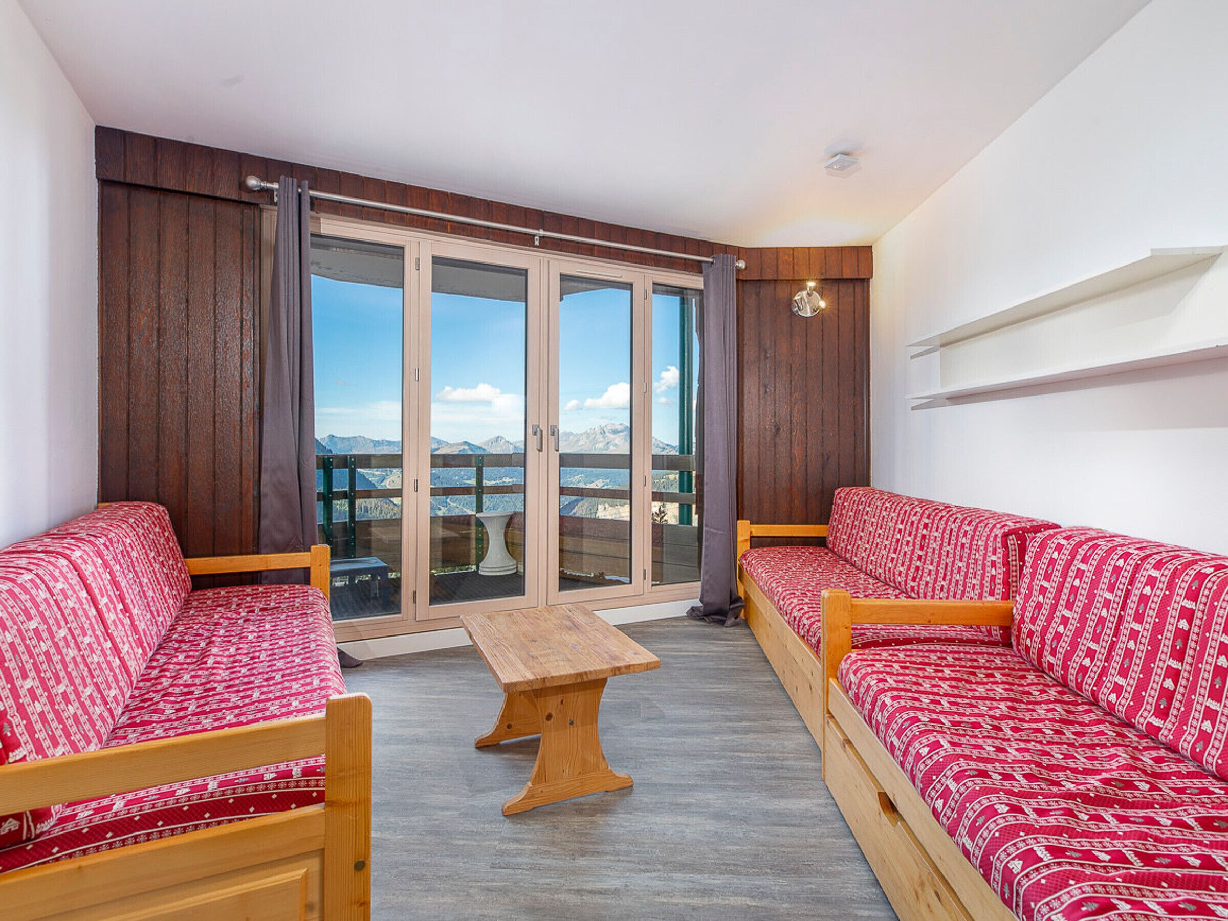 Ein Balkon mit einer hölzernen Bank, zwei rot gemusterten Sofas und Blick auf Berge.
