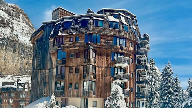 Un edificio de madera de varios pisos con balcones, rodeado de árboles cubiertos de nieve y con una montaña al fondo.