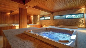 Un espacio de spa con un amplio jacuzzi, techo de madera, paredes de piedra y un ventanal que proporciona luz natural.