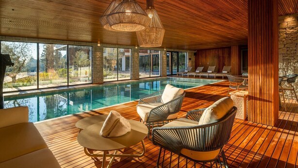 Un área de piscina interior moderna con suelo de madera, sillas de mimbre y ventanas amplias.