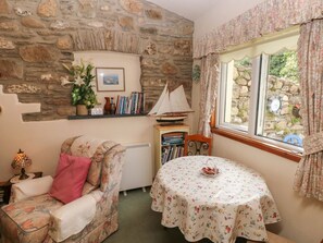 Un cuarto acogedor con pared de piedra, un sillón con estampado floral, una mesita con mantel de flores y una estantería con libros y un barco modelo.