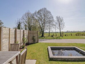 Una terraza de madera con una mesa y sillas, un estanque rectangular y una zona vallada con árboles.