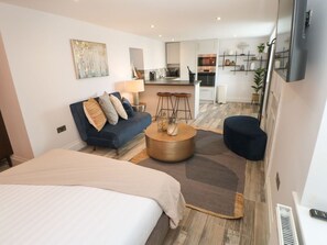 Una habitación de hotel moderna con una cama grande, un sofá, una mesa redonda de centro y una zona de cocina, además de un televisor.