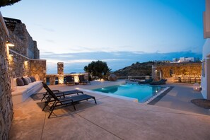 A pool area with lounge chairs, a stone wall, and a view of the sea. - Mykonos Cloud White Villa (Mykonos)