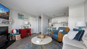 Una sala de estar moderna con un sofá, una mesa de centro y un televisor. Hay una zona de cocina con armarios blancos y una mesa de comedor.