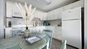 Una cocina con armarios blancos, un refrigerador blanco, una mesa de comedor con sillas y un jarrón con plumas de miscanthus.