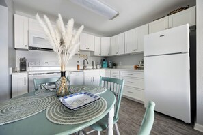 A kitchen with white cabinets, a white refrigerator, a dining table with chairs, and a vase with pampas grass. - Sunset Landing, peaceful water views and front-row seats to some of Ocean City’s stunning sunset (Ocean City)