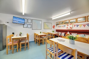 A dining area with wooden tables and chairs, a television mounted on the wall, and a red cushioned bench.