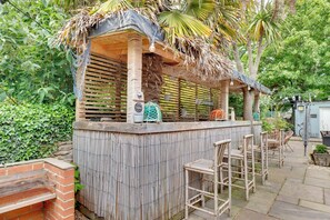 Un bar extérieur en bois, doté d’un toit de chaume, entouré de verdure et agrémenté d’un barbecue en briques.