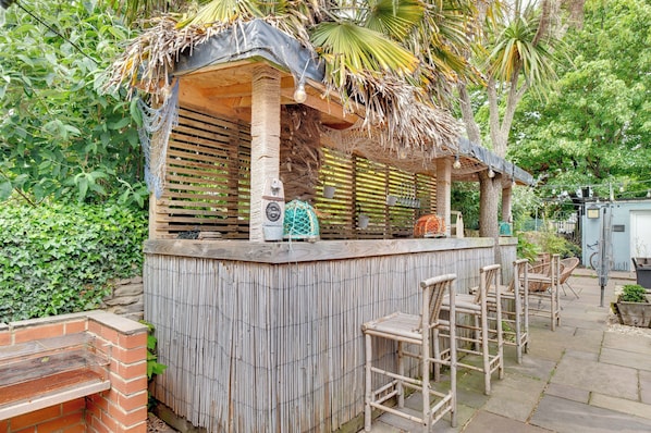 A wooden outdoor bar with a thatched roof, surrounded by greenery and a brick barbecue.