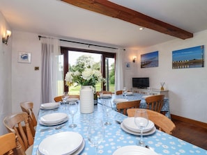 A dining area with a table set for four, chairs, a vase with flowers, and a television on a stand. - 1 Eastdon Cottages, Dawlish Warren (Exeter)