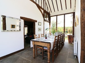 A dining area with a long wooden table set for a meal, flanked by chairs, with a view of greenery outside.