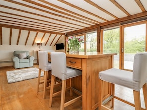 A room with wooden beams, a desk with two chairs, a radiator, a TV, and a window with a view of greenery.