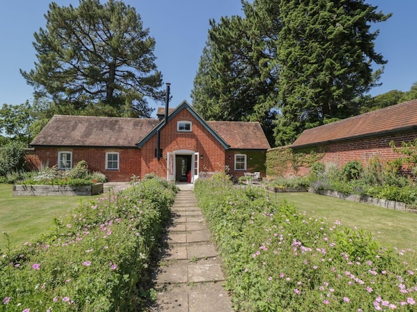A brick house with a front door, a pathway leading to it, and a garden with pink flowers.