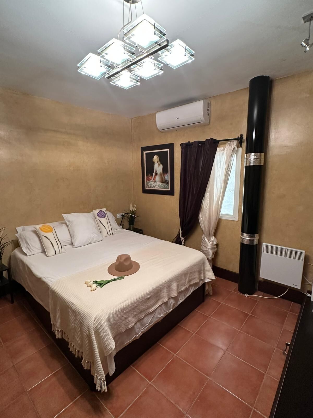 Een slaapkamer met een bed, een plafondlamp, een airconditioning, een raam met gordijnen en een radiator.