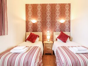 Une chambre d’hôtel avec deux lits, chacun agrémenté d’un oreiller rouge et de draps blancs, une table de chevet avec une lampe et un papier peint à motifs.