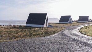 A gravel road leading to three white buildings with dark roofs, situated in a rural landscape with a body of water and hills in the background. - Cabin 2 Min Larach (Gortahork)