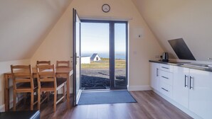 A room with a dining table and chairs, a kitchenette, a large sliding glass door leading to a view, and a clock on the wall. - Cabin 2 Min Larach (Gortahork)
