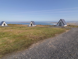 Three small, white, triangular-roofed cabins with a gravel path leading to them, set against a coastal backdrop. - Cabin 1 Machaire Rabhartaigh (Gortahork)