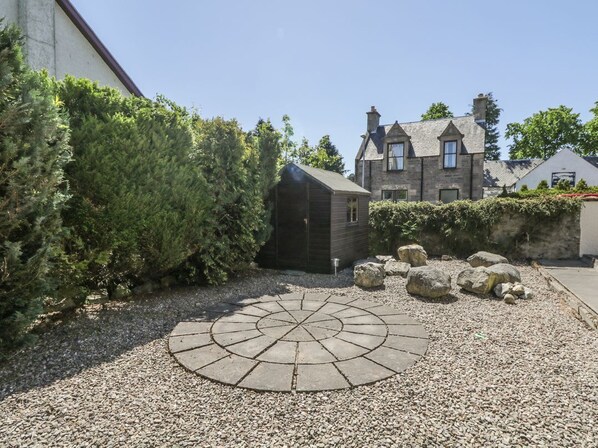 A courtyard with a circular stone pattern, a small wooden structure, and a stone wall with a house in the background. - Dalnahaven: Your Dog-Friendly Retreat (Grantown-on-Spey)